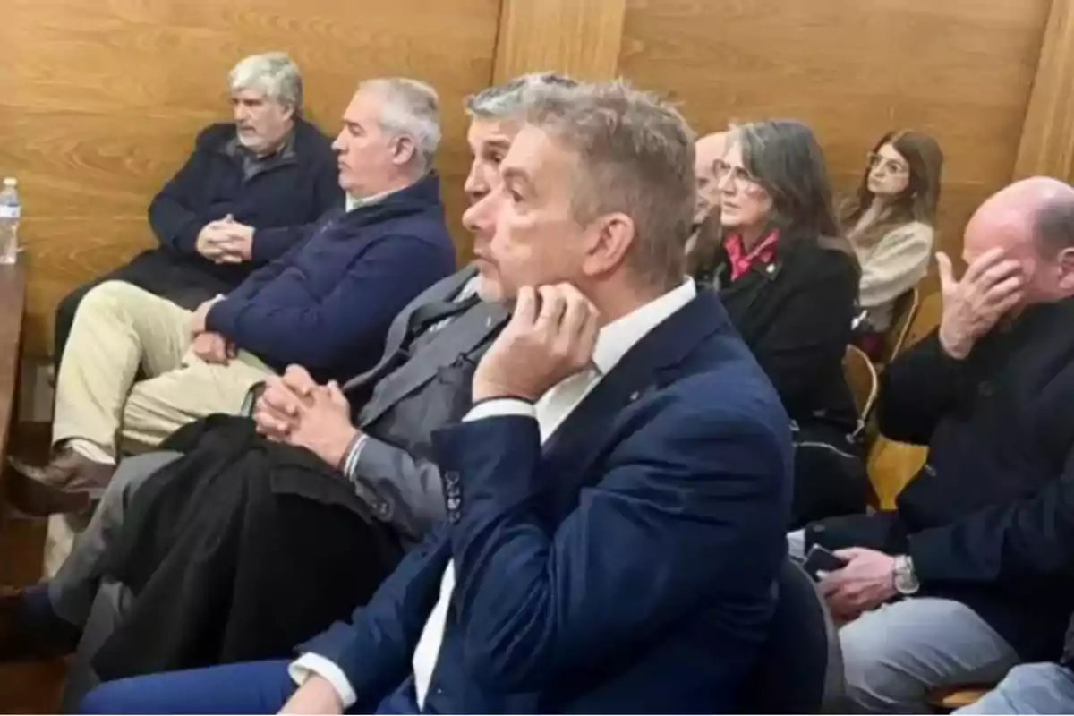 Several people sitting in a room with serious and formal expressions, some with their hands clasped and one person covering their face with their hand.