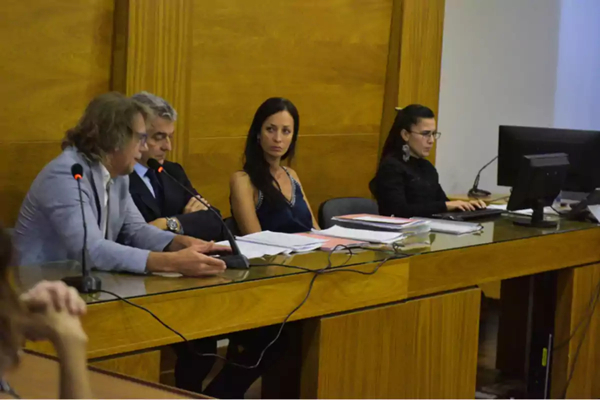 People seated at a wooden table in a courtroom, with microphones, documents, and a computer, participating in a judicial proceeding. People seated at a wooden table in a courtroom, with microphones, documents, and a computer, participating in a judicial proceeding.
