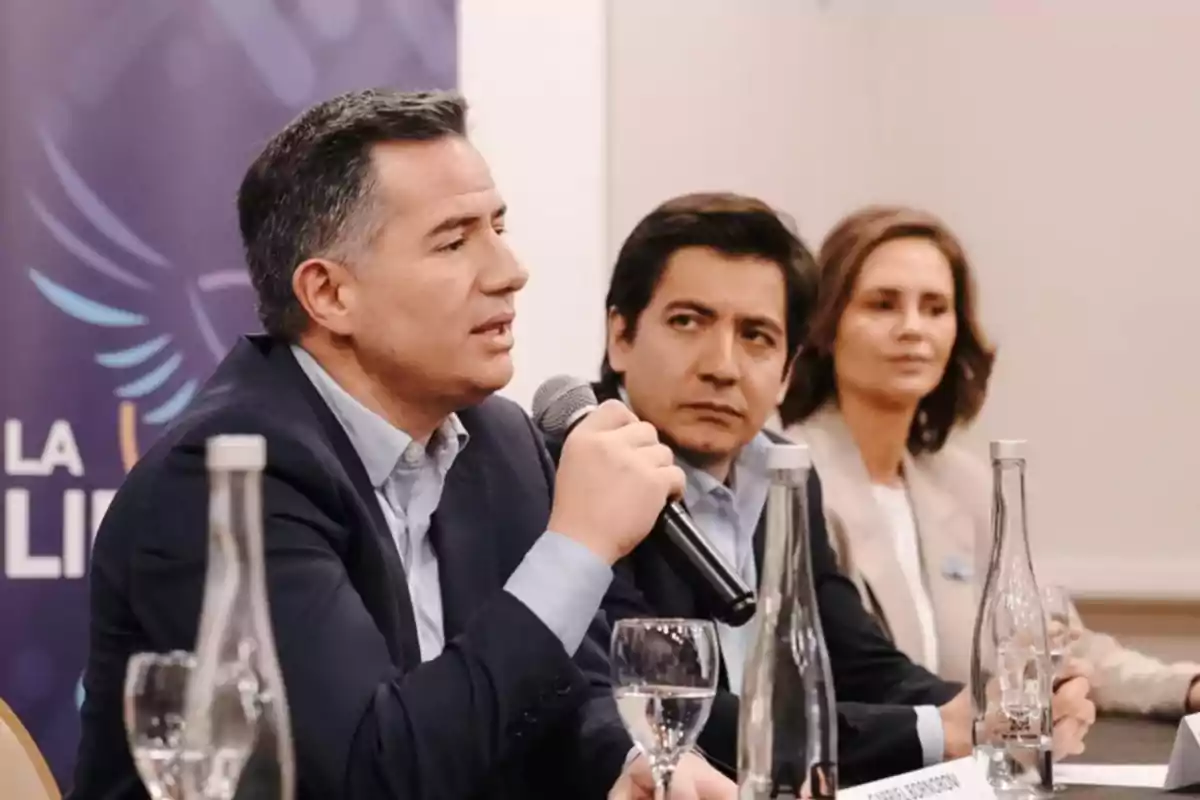 Three people sitting at a table during a conference while one of them speaks into a microphone Three people sitting at a table during a conference while one of them speaks into a microphone