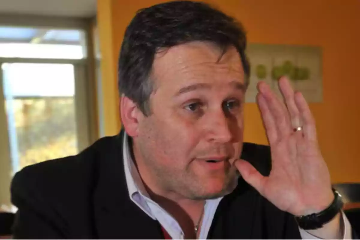 Middle-aged man with dark hair and a black jacket gesturing with his hand near his face in a well-lit indoor setting Middle-aged man with dark hair and a black jacket gesturing with his hand near his face in a well-lit indoor setting