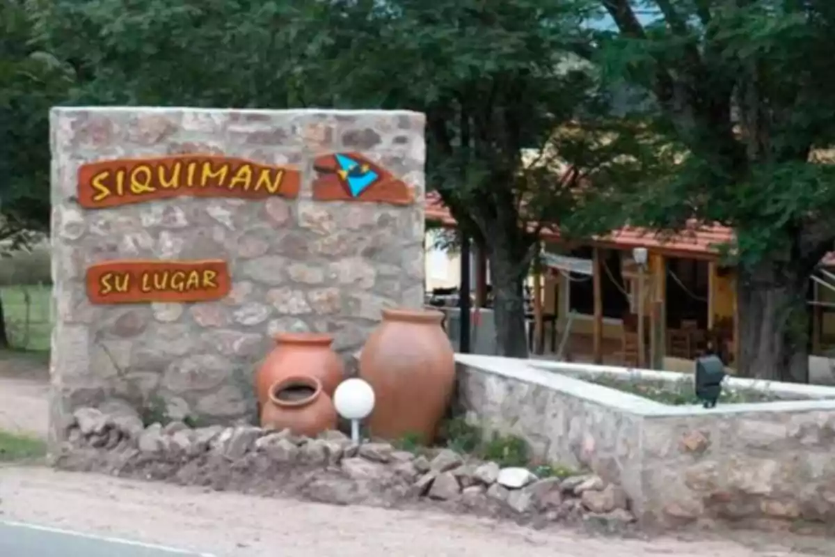 Cartel de bienvenida de piedra con la palabra Siquiman y la frase su lugar acompañado de dos grandes vasijas de barro y árboles de fondo Cartel de bienvenida de piedra con la palabra Siquiman y la frase su lugar acompañado de dos grandes vasijas de barro y árboles de fondo