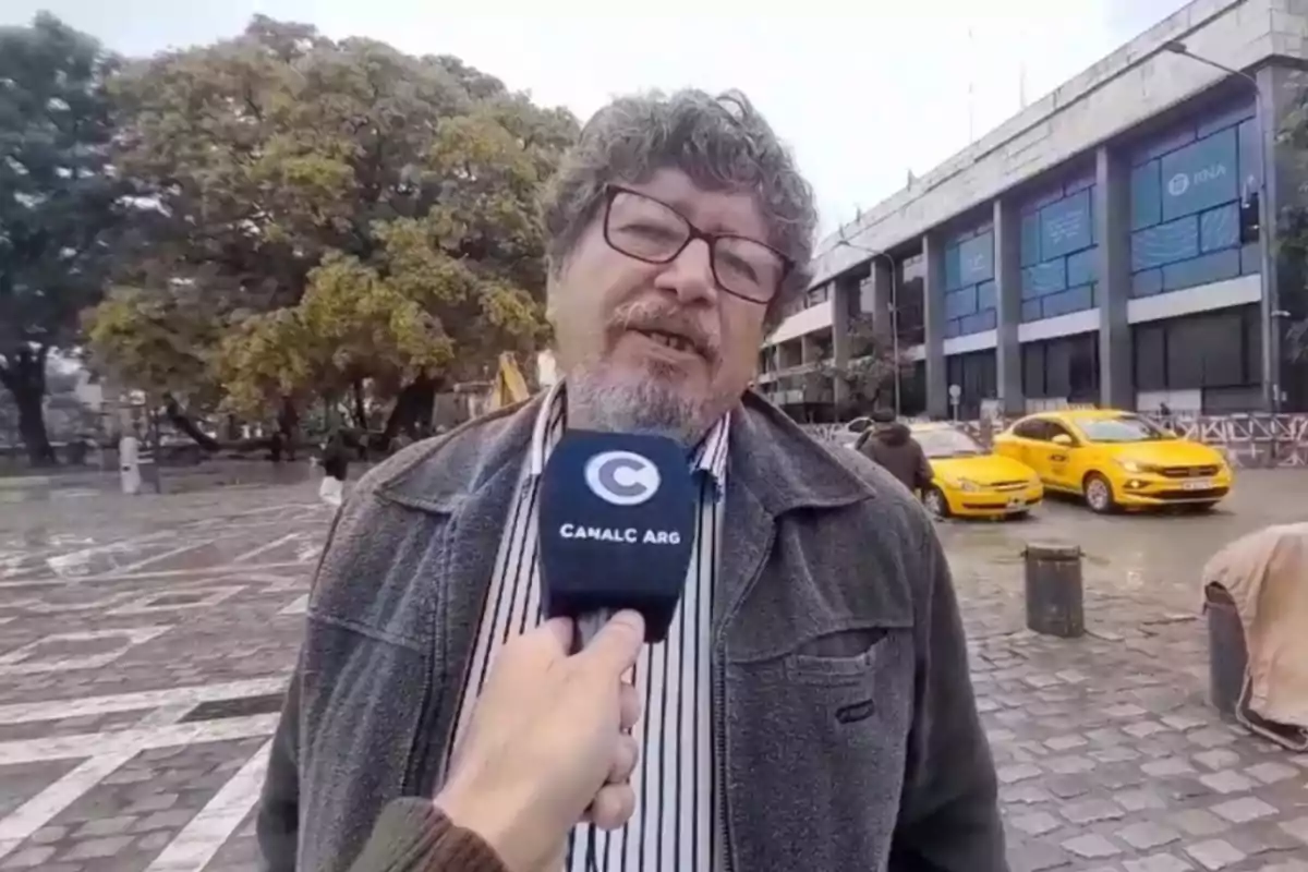 Un hombre con gafas y barba es entrevistado en la calle por un canal de televisión mientras dos taxis amarillos pasan por detrás y se observa un edificio moderno y árboles al fondo Un hombre con gafas y barba es entrevistado en la calle por un canal de televisión mientras dos taxis amarillos pasan por detrás y se observa un edificio moderno y árboles al fondo