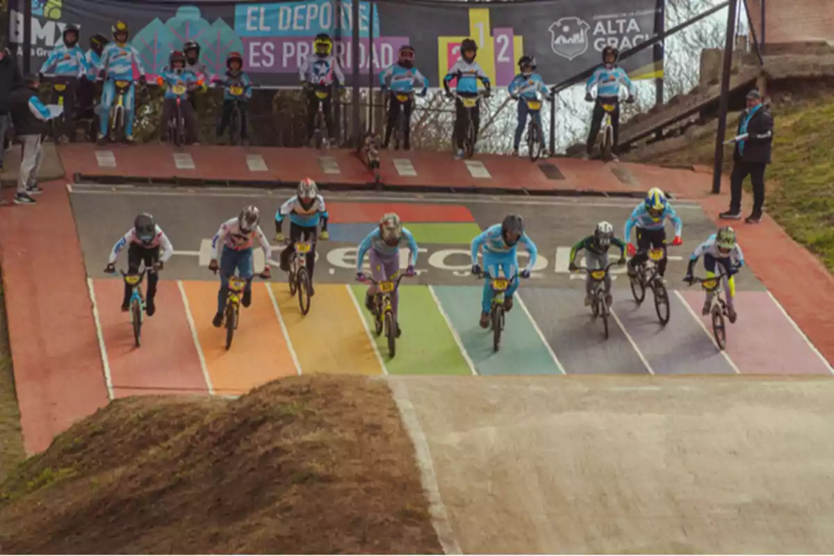 Varios ciclistas compiten en una carrera de BMX saliendo desde la línea de inicio en una pista con franjas de colores mientras otros participantes observan desde el fondo