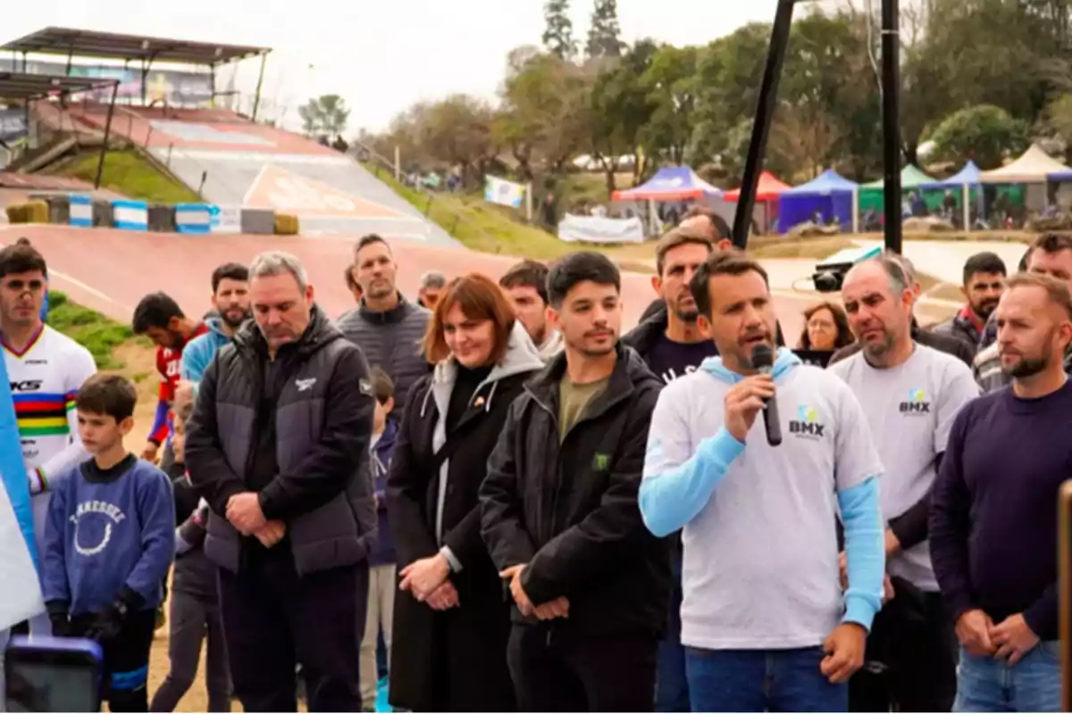 A group of people gathered outdoors at a BMX track while a man with a microphone speaks to the audience. A group of people gathered outdoors at a BMX track while a man with a microphone speaks to the audience.