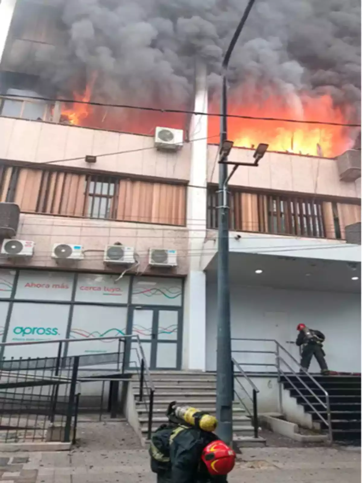 Edificio en llamas con humo negro saliendo de las ventanas y dos bomberos en la entrada preparados para intervenir Edificio en llamas con humo negro saliendo de las ventanas y dos bomberos en la entrada preparados para intervenir