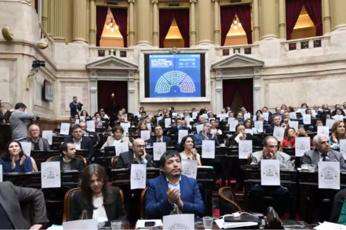 Parliamentary session with numerous legislators seated at their desks inside a chamber with classical architecture and a large screen in the background displaying voting results. Parliamentary session with numerous legislators seated at their desks inside a chamber with classical architecture and a large screen in the background displaying voting results.