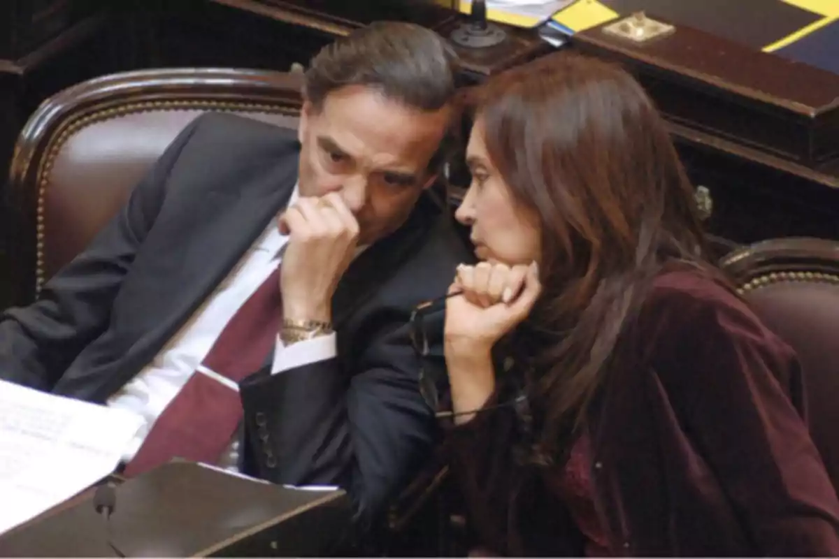 Two people sitting in a parliamentary chamber speak in low voices while covering their mouths with their hands.
