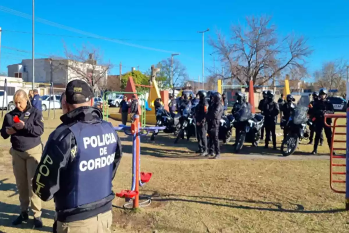 Several police officers from Córdoba are gathered in an outdoor park alongside motorcycles and riot control equipment on a sunny day.