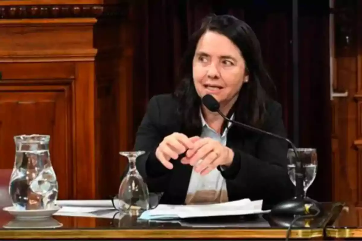 Dark-haired woman sitting at a table with a microphone, papers, and glasses of water, speaking in a formal wooden setting