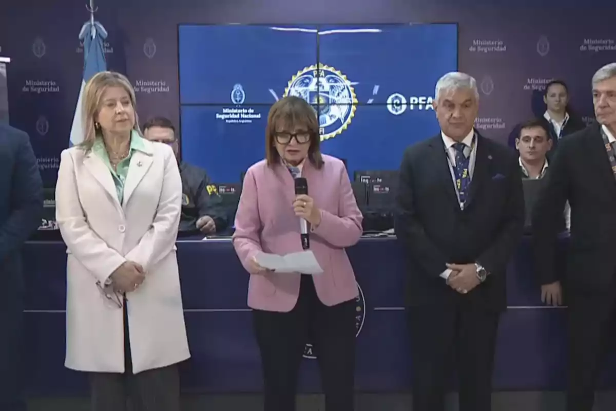 Several people standing at an official event in a Ministry of Security room, with a woman in the center speaking into a microphone and blue screens in the background displaying the institutional logo.