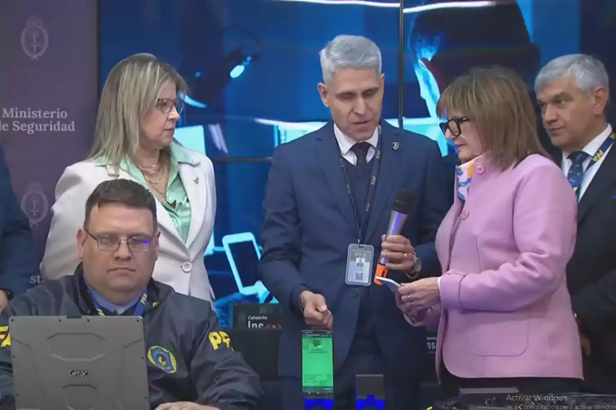 Varias personas con credenciales y uniforme de la policía federal argentina están reunidas en una oficina con computadoras y dispositivos electrónicos, mientras una mujer sostiene un micrófono y conversa con un hombre de cabello canoso