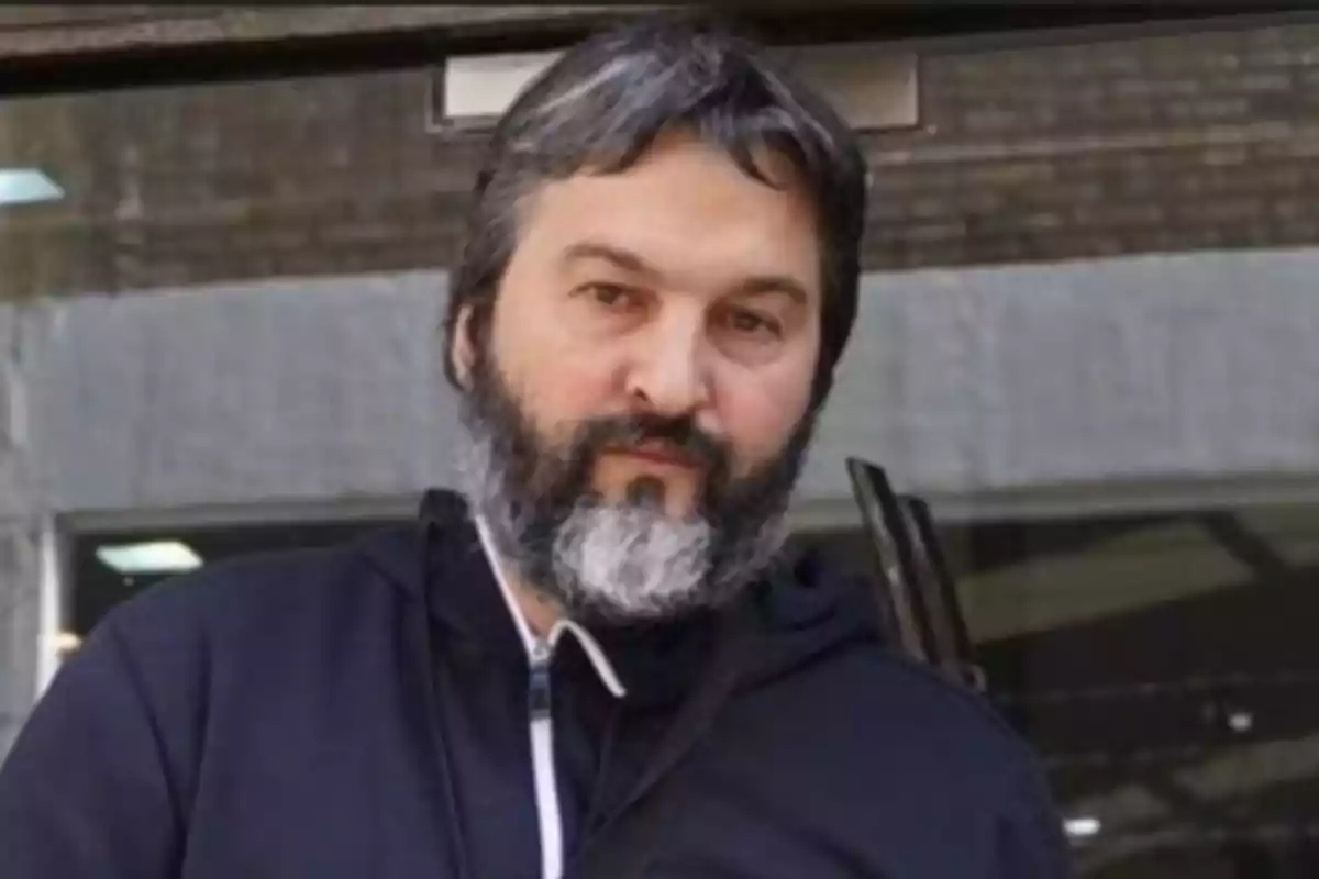 Hombre de cabello y barba canosa vistiendo una chaqueta oscura frente a un edificio