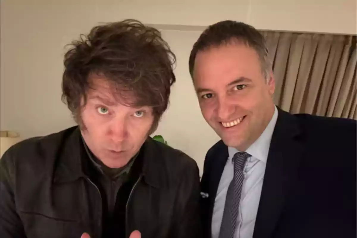 Dos hombres posan juntos para una selfie en una habitación iluminada, uno lleva chaqueta de cuero y el otro viste traje y corbata