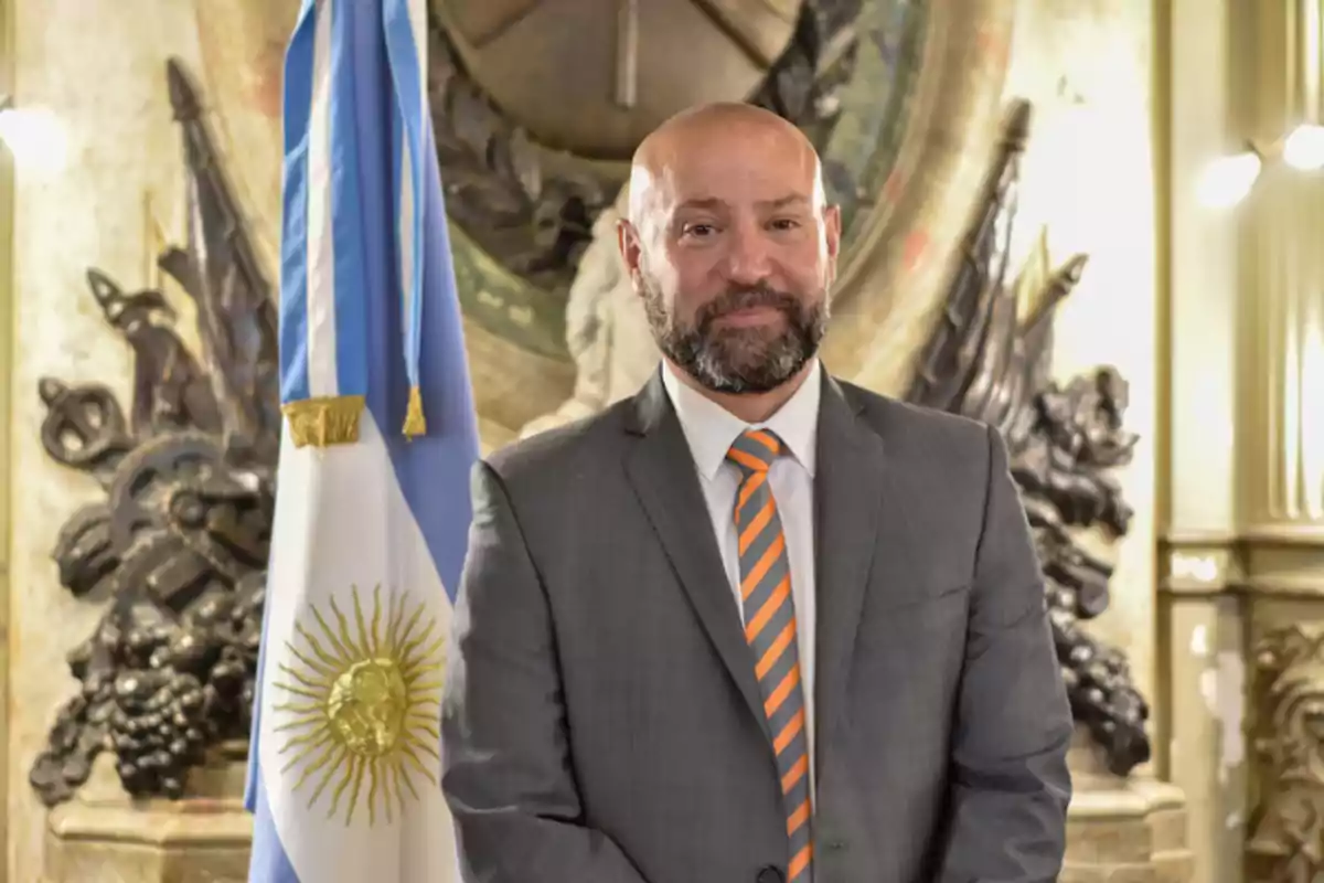 Man in a gray suit with an orange and gray striped tie posing in front of an Argentine flag in a formal setting