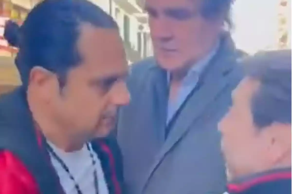 Tres hombres conversan de cerca en una calle, uno de ellos lleva chaqueta roja y otro viste saco gris.
