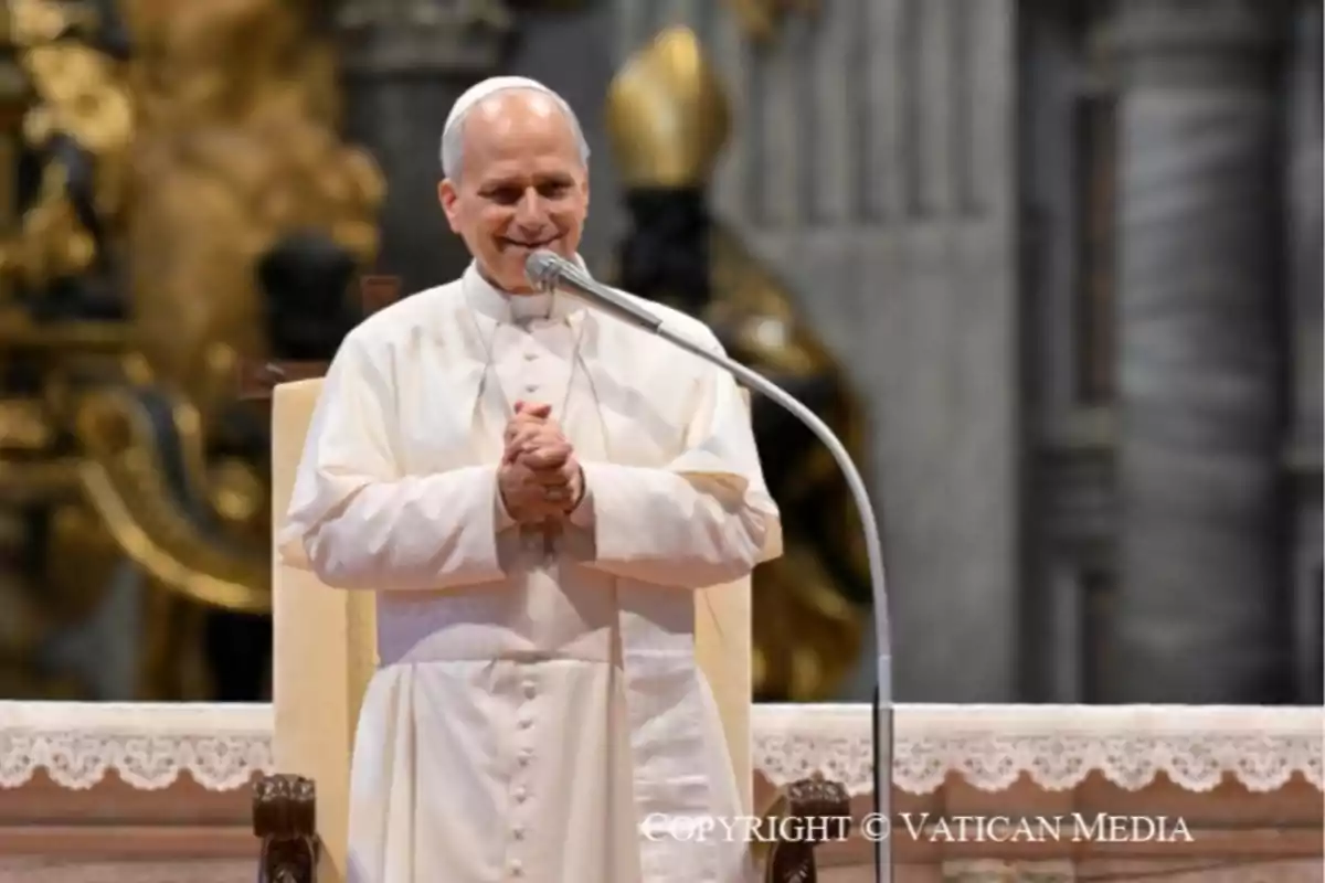 Hombre vestido con sotana blanca y solideo hablando sonriente frente a un micrófono en un entorno religioso Hombre vestido con sotana blanca y solideo hablando sonriente frente a un micrófono en un entorno religioso