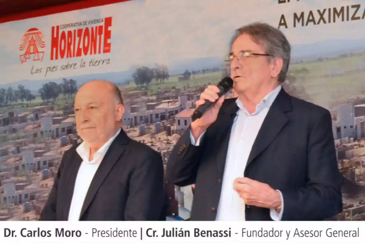 Two men in suits are standing in front of a Horizonte housing cooperative sign; one of them is holding a microphone while the other listens alertly.