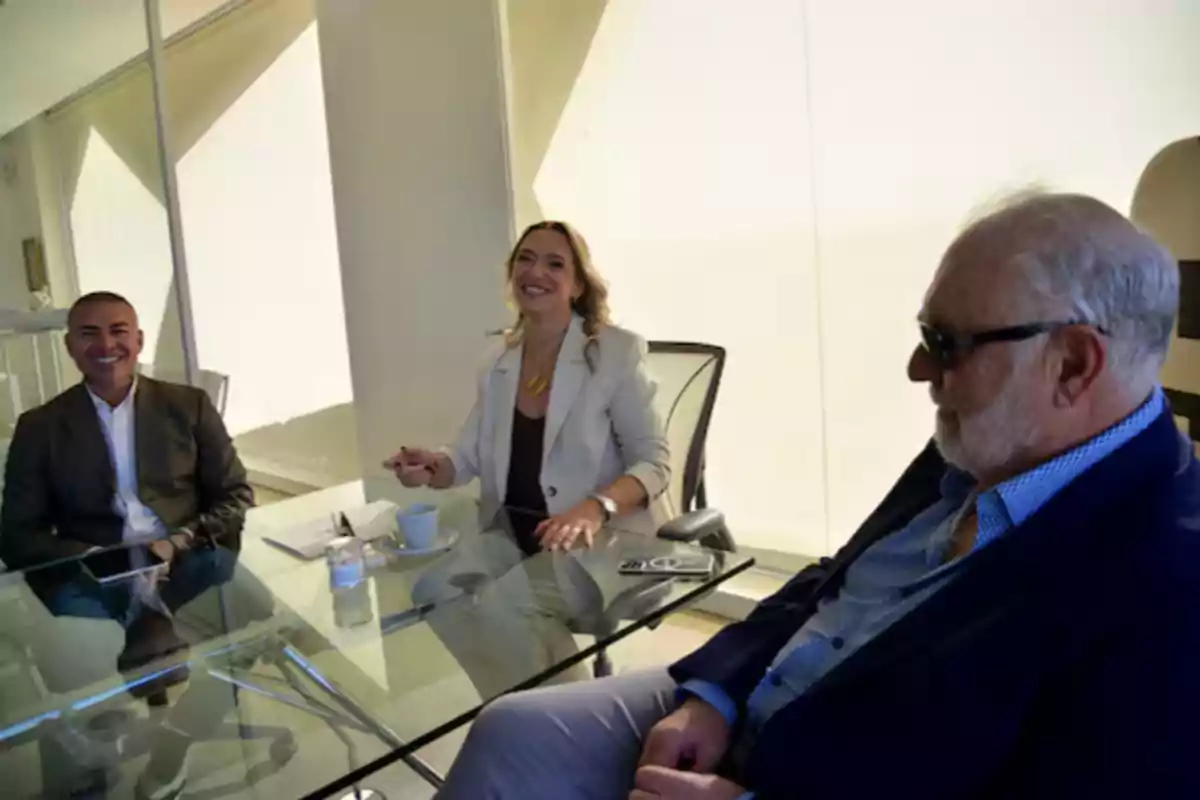 Three people smile while talking, seated around a glass table in a brightly lit office. Three people smile while talking, seated around a glass table in a brightly lit office.