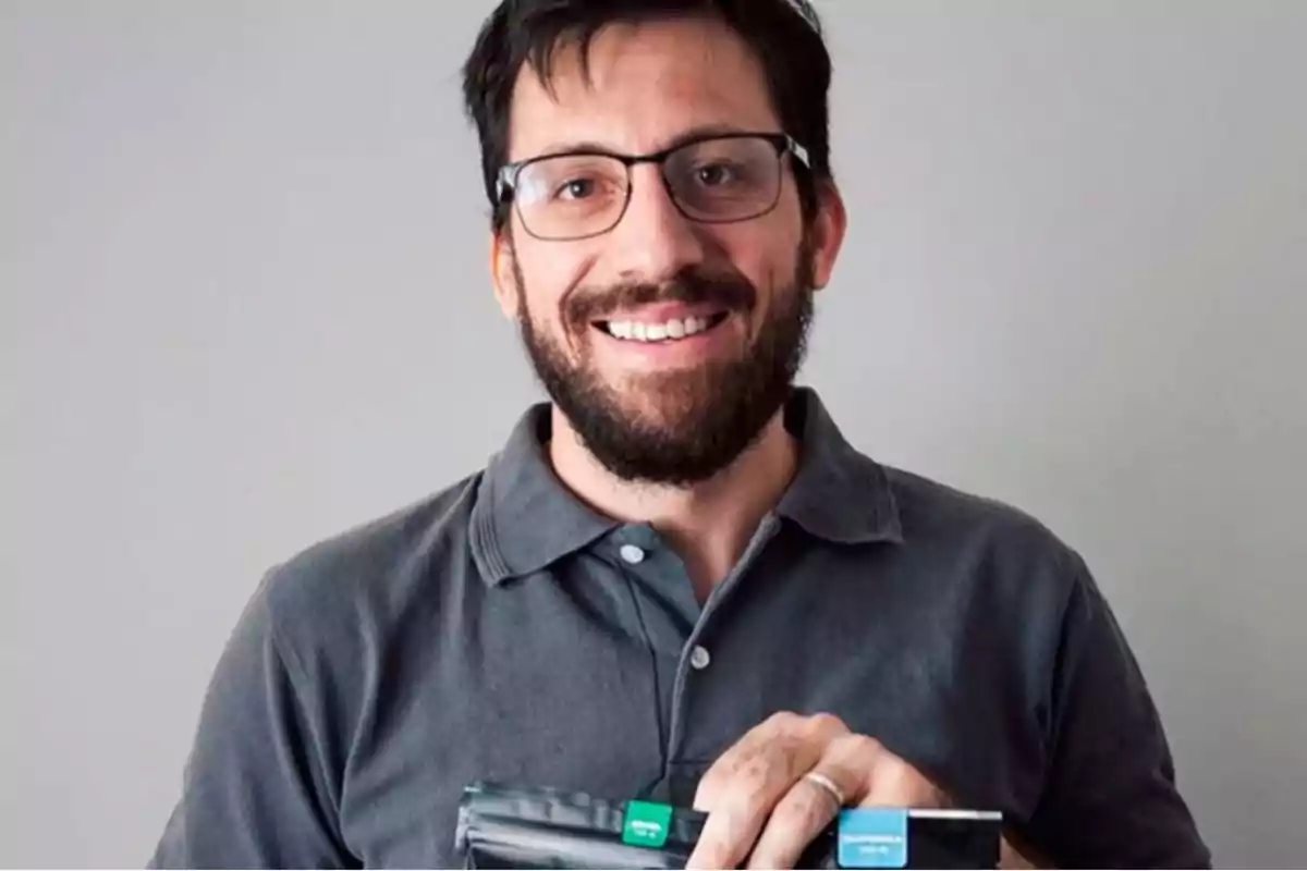 Man with a beard and glasses smiling while holding a black bag with colorful tags against a gray background.