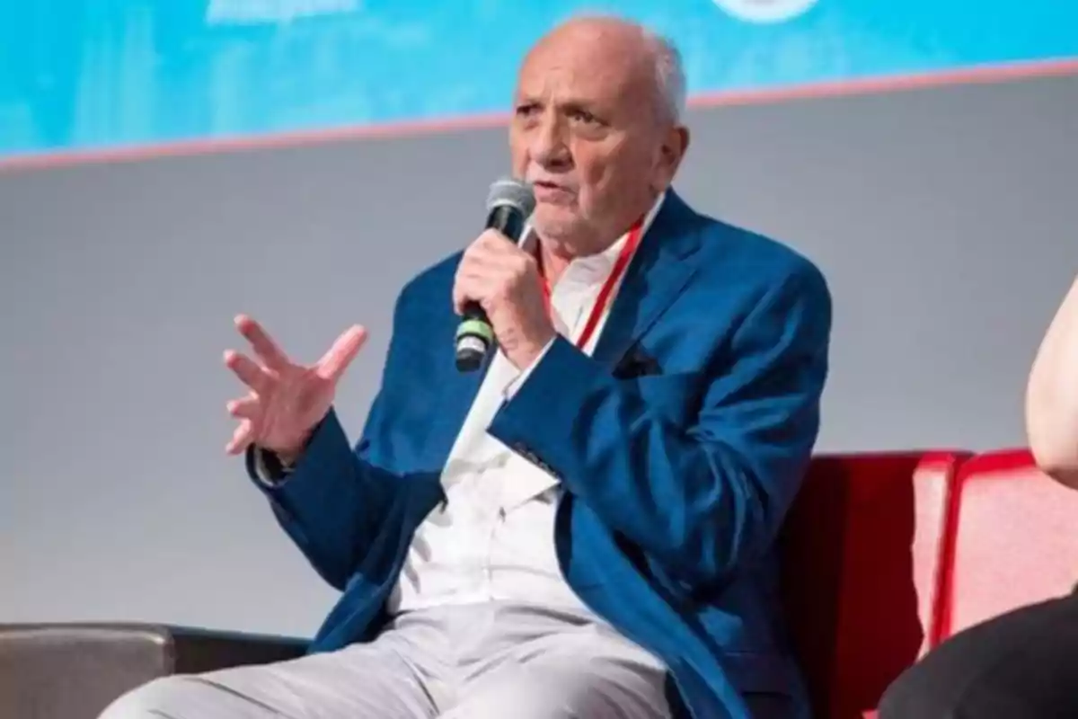 Older man in a blue jacket speaking into a microphone while sitting in a red armchair Older man in a blue jacket speaking into a microphone while sitting in a red armchair