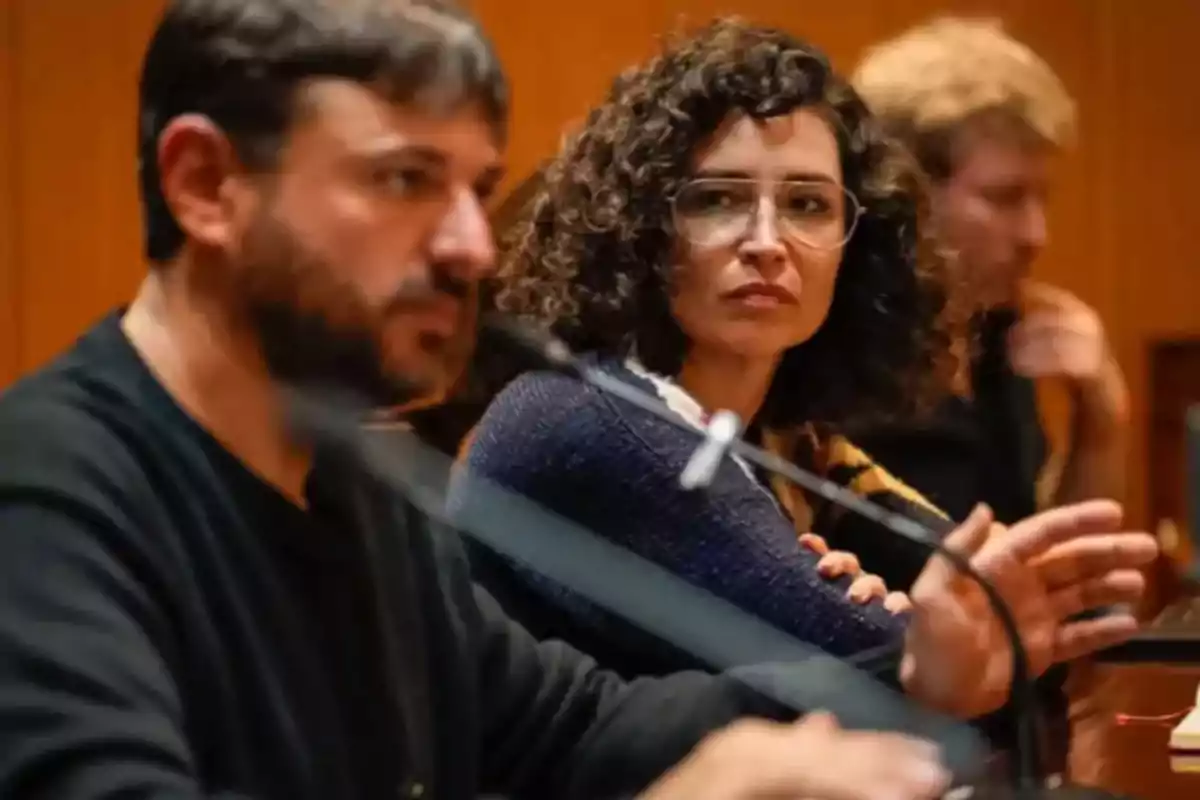 Three people sitting at a table engaged in a conversation, a woman with glasses and curly hair alertly looks at the man who is speaking in front of a microphone.