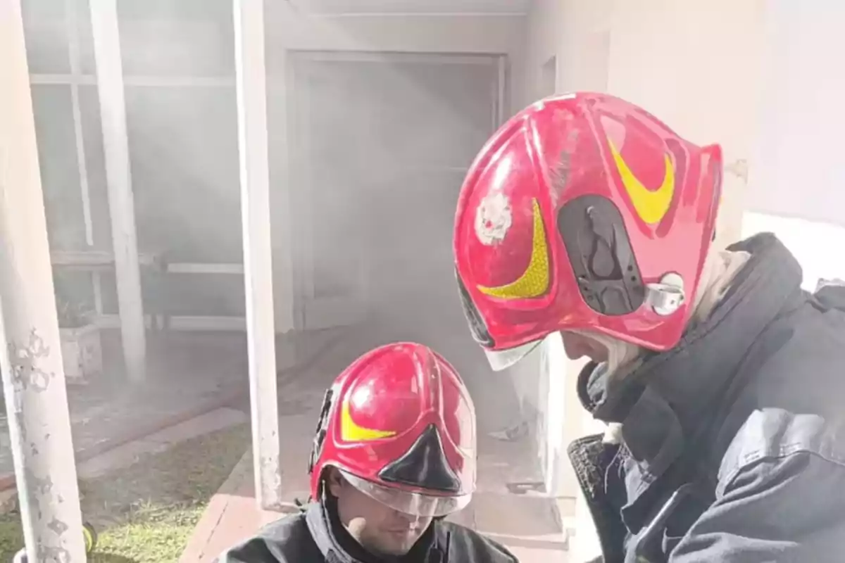 Two firefighters wearing red helmets and protective gear are in a smoky building; one of them appears to be assisting the other. Two firefighters wearing red helmets and protective gear are in a smoky building; one of them appears to be assisting the other.