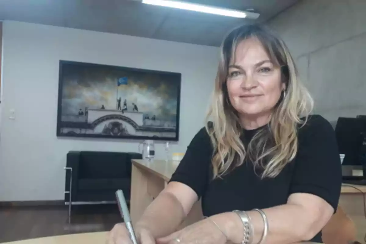 Blonde-haired woman sitting in an office writing, with a painting on the wall in the background