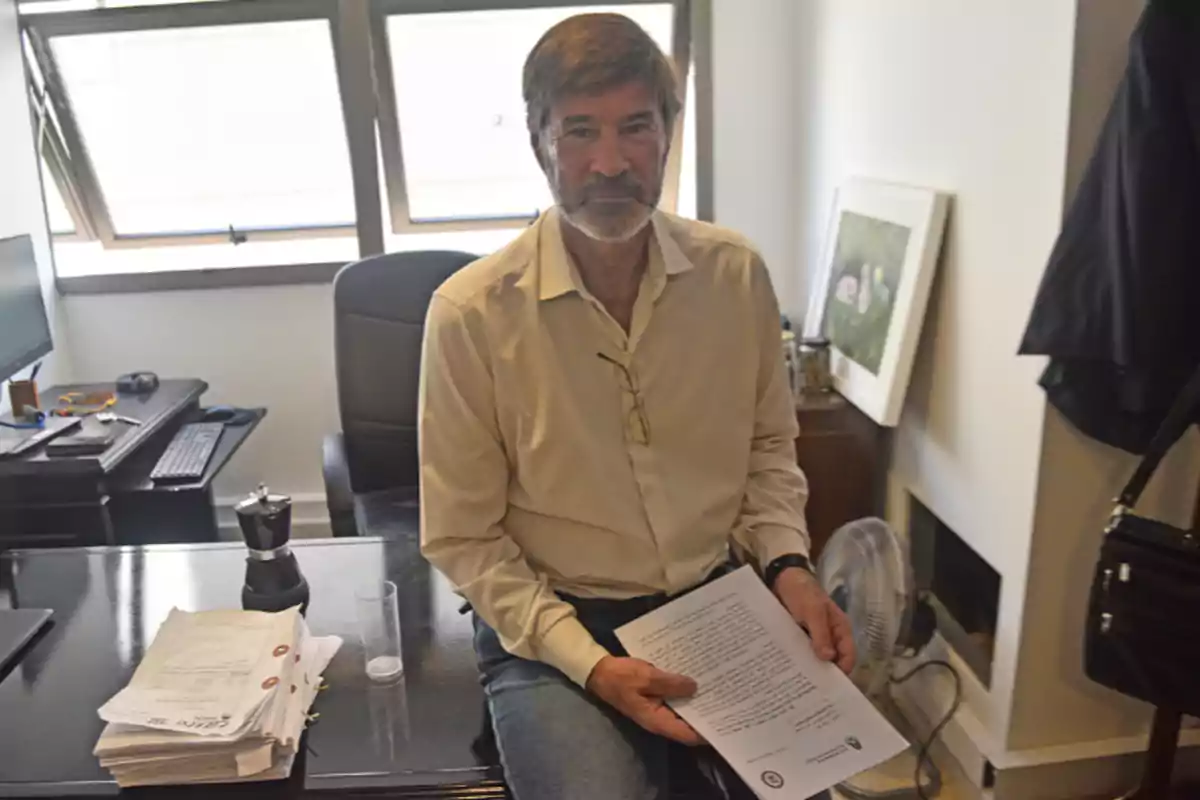 Gray-haired, bearded man sitting in an office holding some papers, with a desk and a computer in the background Gray-haired, bearded man sitting in an office holding some papers, with a desk and a computer in the background