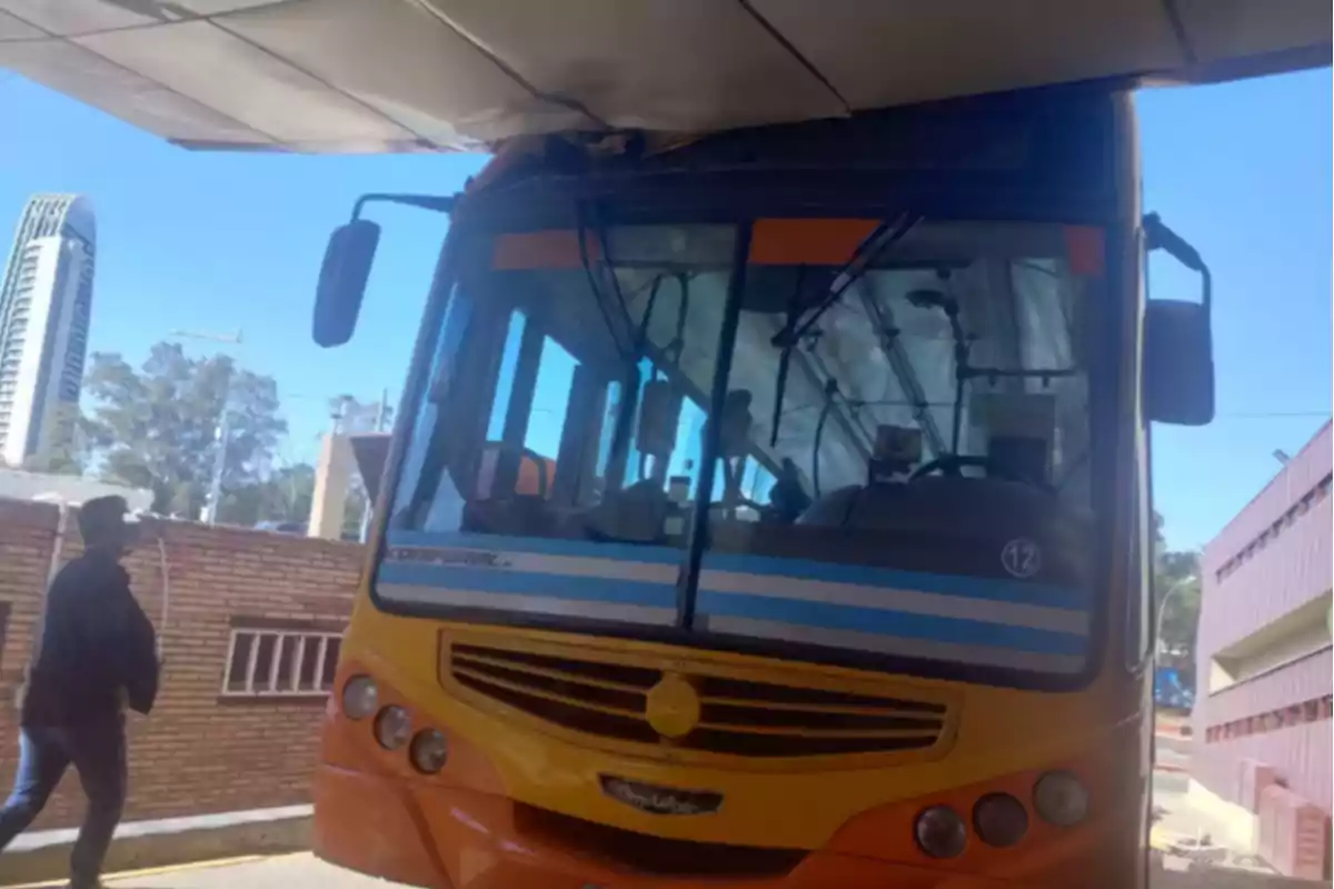 Yellow bus stuck under a roof that's too low at a terminal, with a person walking nearby.