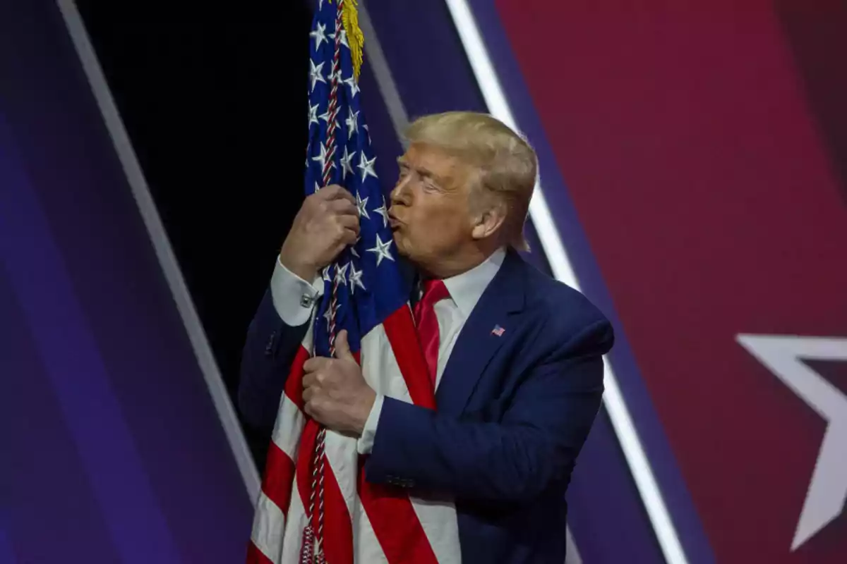 Man in a blue suit hugging and kissing a United States flag on a stage with a red and purple background