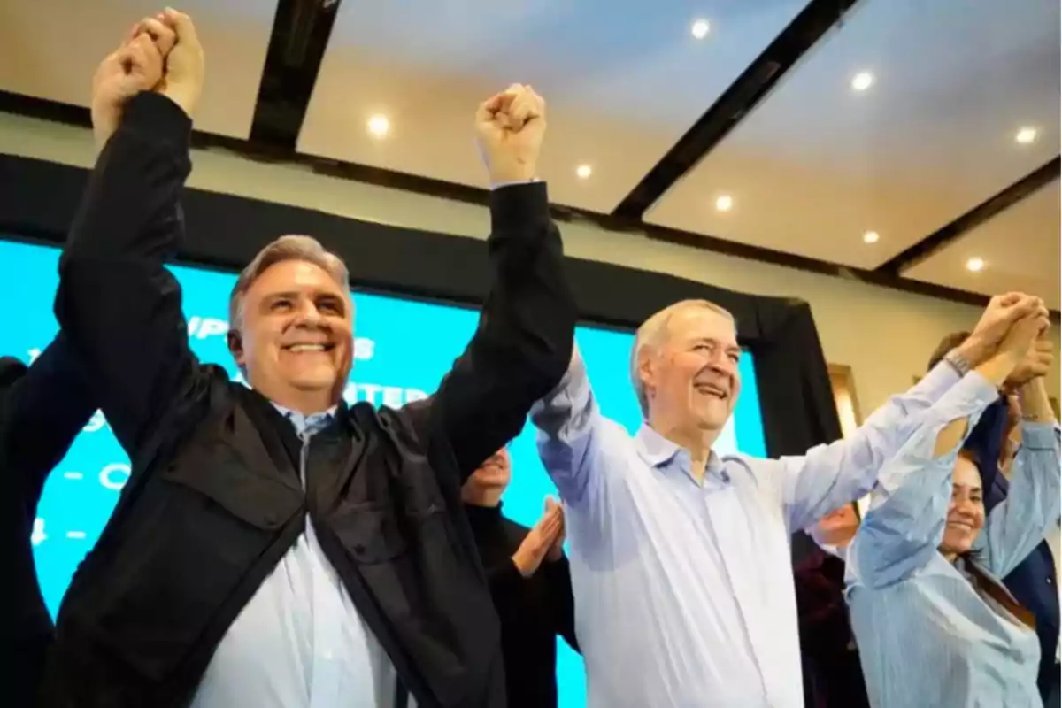 Two smiling men raise their interlocked arms in celebration at an indoor event, accompanied by other people in the background. Two smiling men raise their interlocked arms in celebration at an indoor event, accompanied by other people in the background.