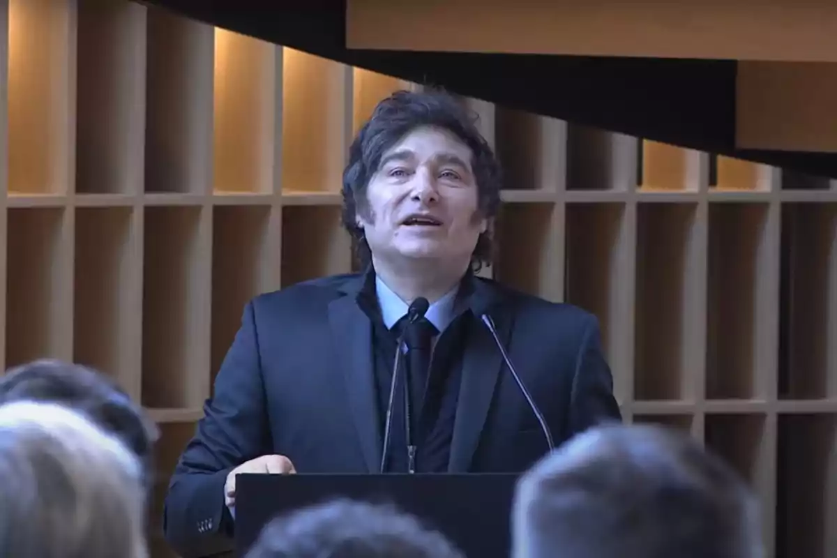 Dark-haired man in a black suit speaking at a podium in front of an audience in a modern auditorium