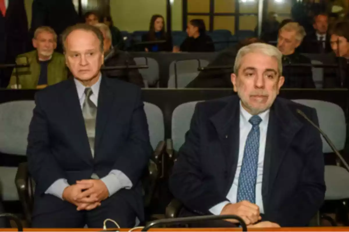 Two men in suits sitting in a room with several people in the background Two men in suits sitting in a room with several people in the background