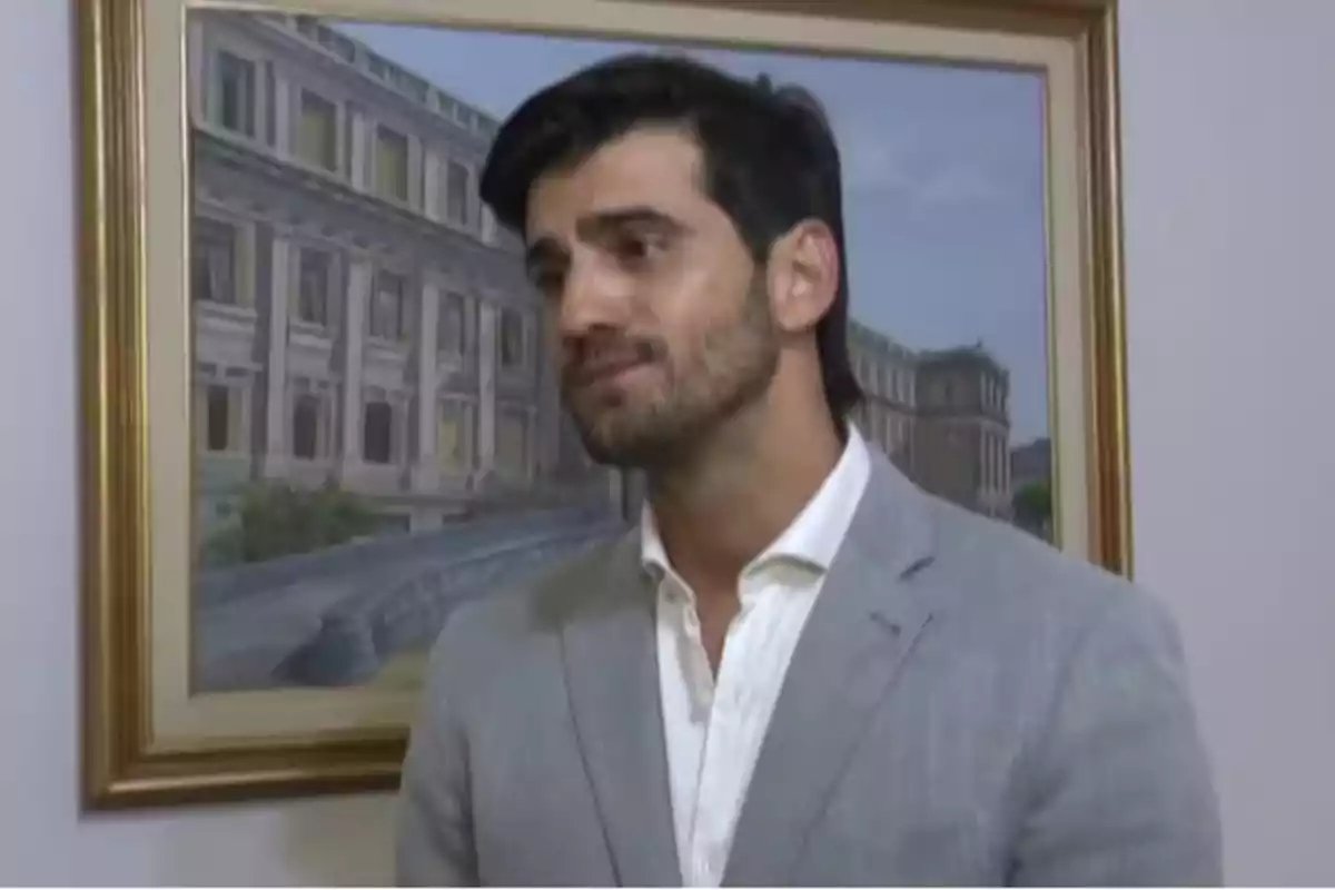 Hombre de cabello oscuro y barba corta vistiendo un saco gris claro y camisa blanca de pie frente a un cuadro de un edificio antiguo Hombre de cabello oscuro y barba corta vistiendo un saco gris claro y camisa blanca de pie frente a un cuadro de un edificio antiguo