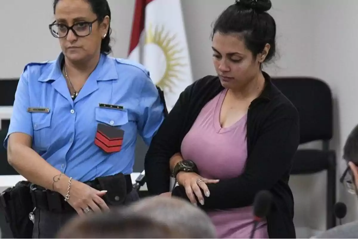 A female police officer escorts another woman with a serious expression in a room where more people are present.