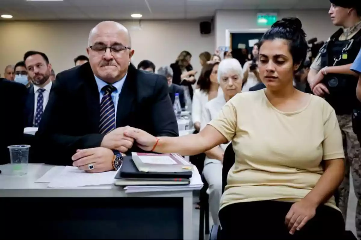 A man in a suit holds the hand of a woman with a serious expression in a room full of people, both seated in front of a table with documents and books. A man in a suit holds the hand of a woman with a serious expression in a room full of people, both seated in front of a table with documents and books.