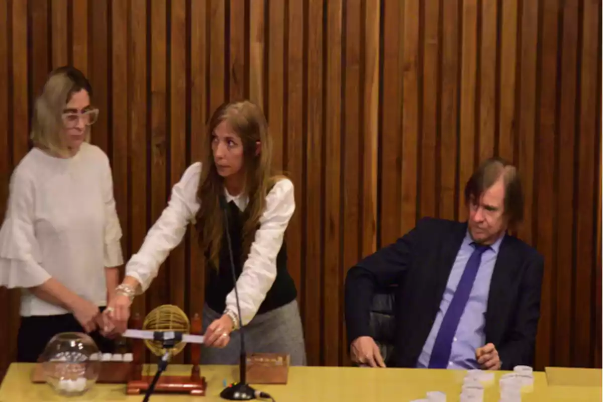 Tres personas en una mesa de madera, dos mujeres están manipulando una tómbola y un hombre con traje está sentado observando