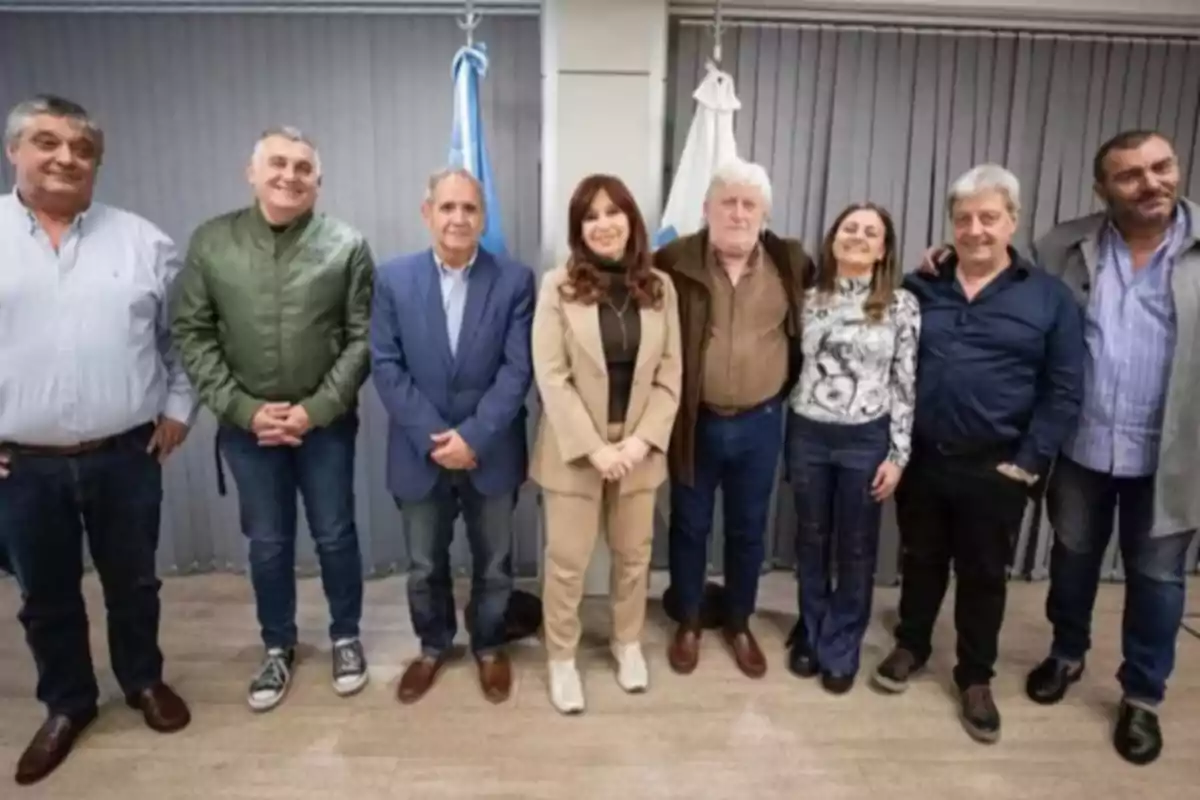 Eight people stand in a row inside an office with vertical blinds and two flags in the background. Eight people stand in a row inside an office with vertical blinds and two flags in the background.