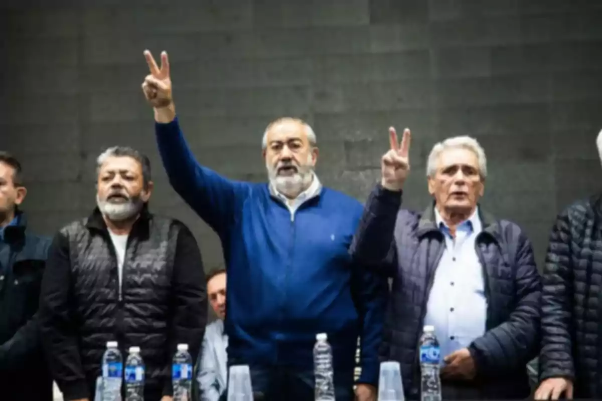 Five men are standing at a table, two of them are raising their hands making the victory sign, there are water bottles and glasses on the table, and they are all wearing warm clothing.
