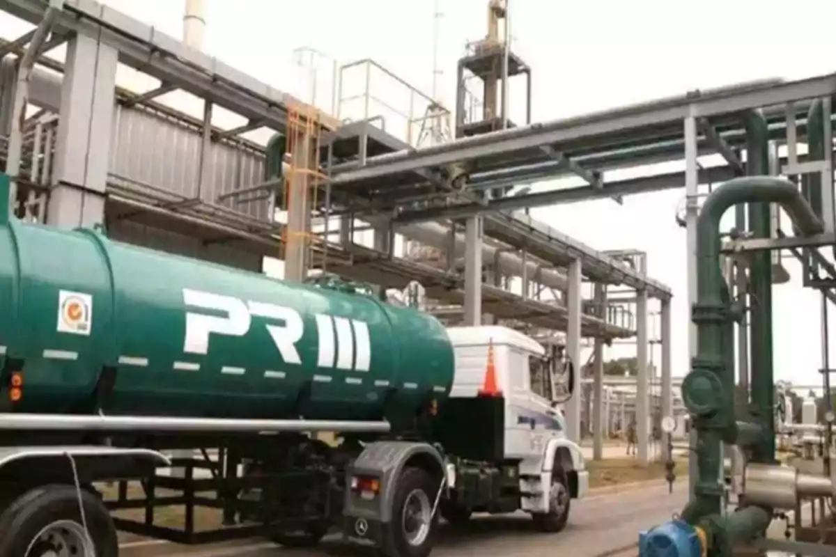 Green tanker truck parked at an industrial plant surrounded by pipes and metal structures