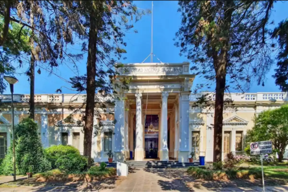 Main facade of a historic building surrounded by trees and gardens, with white columns and a sign at the top Main facade of a historic building surrounded by trees and gardens, with white columns and a sign at the top
