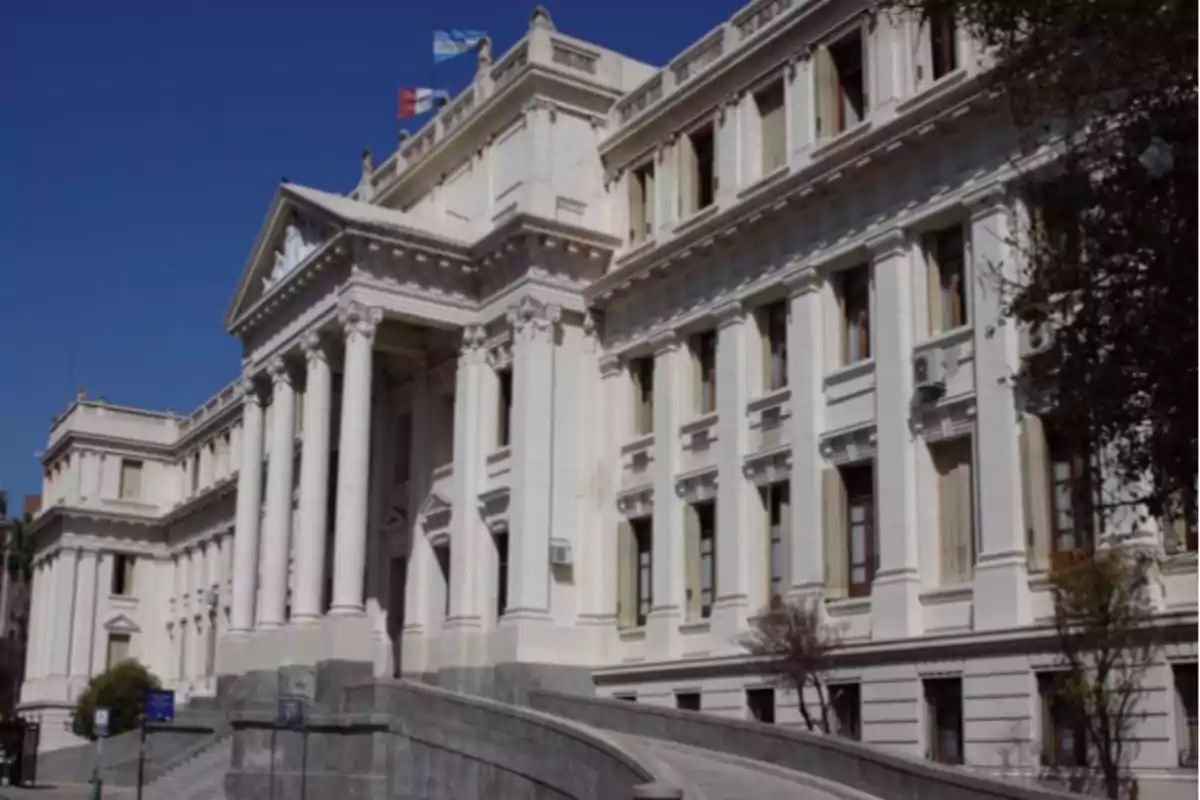 Facade of a historic building with classical architecture, columns, and flags on the roof under a clear sky