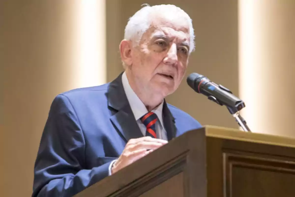 Older man with gray hair wearing a blue suit and tie, speaking at a podium in front of a microphone Older man with gray hair wearing a blue suit and tie, speaking at a podium in front of a microphone