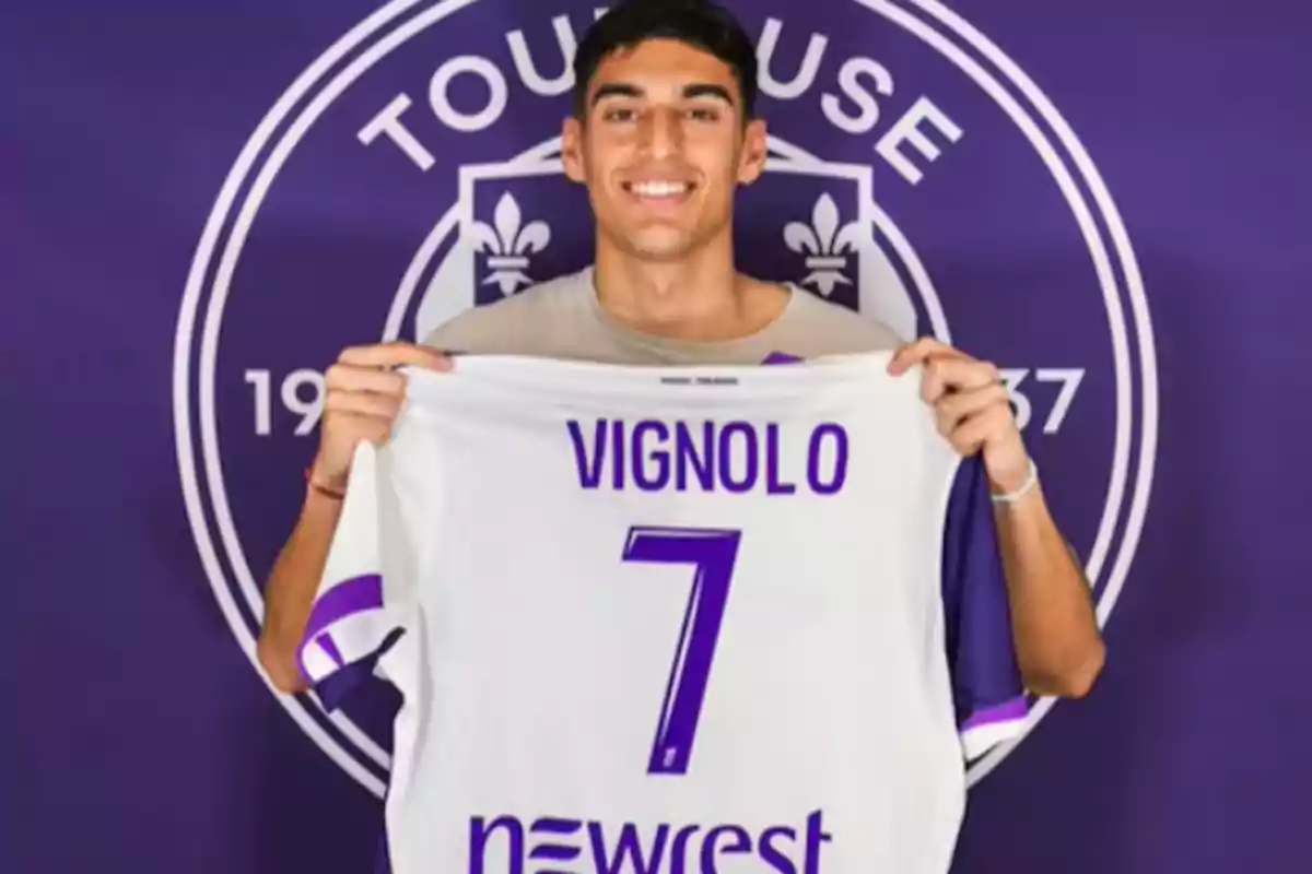 A smiling young man holds a white soccer jersey with purple details that has the name Vignolo and the number 7 in front of a background featuring the Toulouse crest.