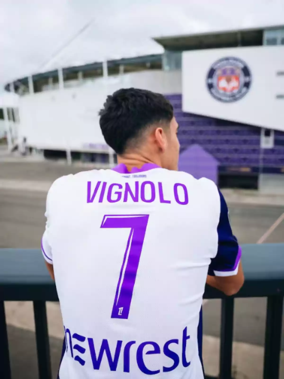 Person seen from behind wearing a white and purple soccer jersey with the name Vignolo and the number 7, looking toward a stadium. Person seen from behind wearing a white and purple soccer jersey with the name Vignolo and the number 7, looking toward a stadium.