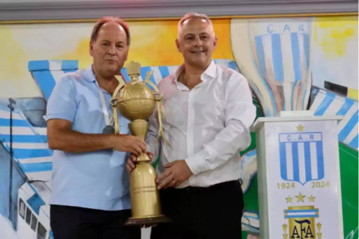 Two men pose smiling, holding a golden trophy in front of a mural with light blue and white colors and a podium with the crest of an Argentine soccer club. Two men pose smiling, holding a golden trophy in front of a mural with light blue and white colors and a podium with the crest of an Argentine soccer club.