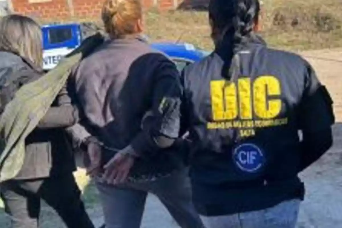 Tres personas de espaldas, una de ellas esposada y escoltada por dos agentes, una de las cuales lleva una chaqueta con las siglas DIC en amarillo.