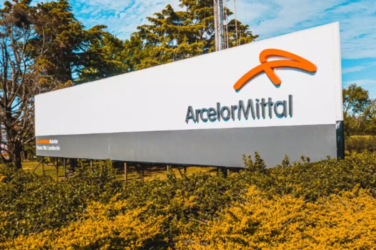 Letrero de la empresa ArcelorMittal rodeado de vegetación y árboles bajo un cielo azul Letrero de la empresa ArcelorMittal rodeado de vegetación y árboles bajo un cielo azul