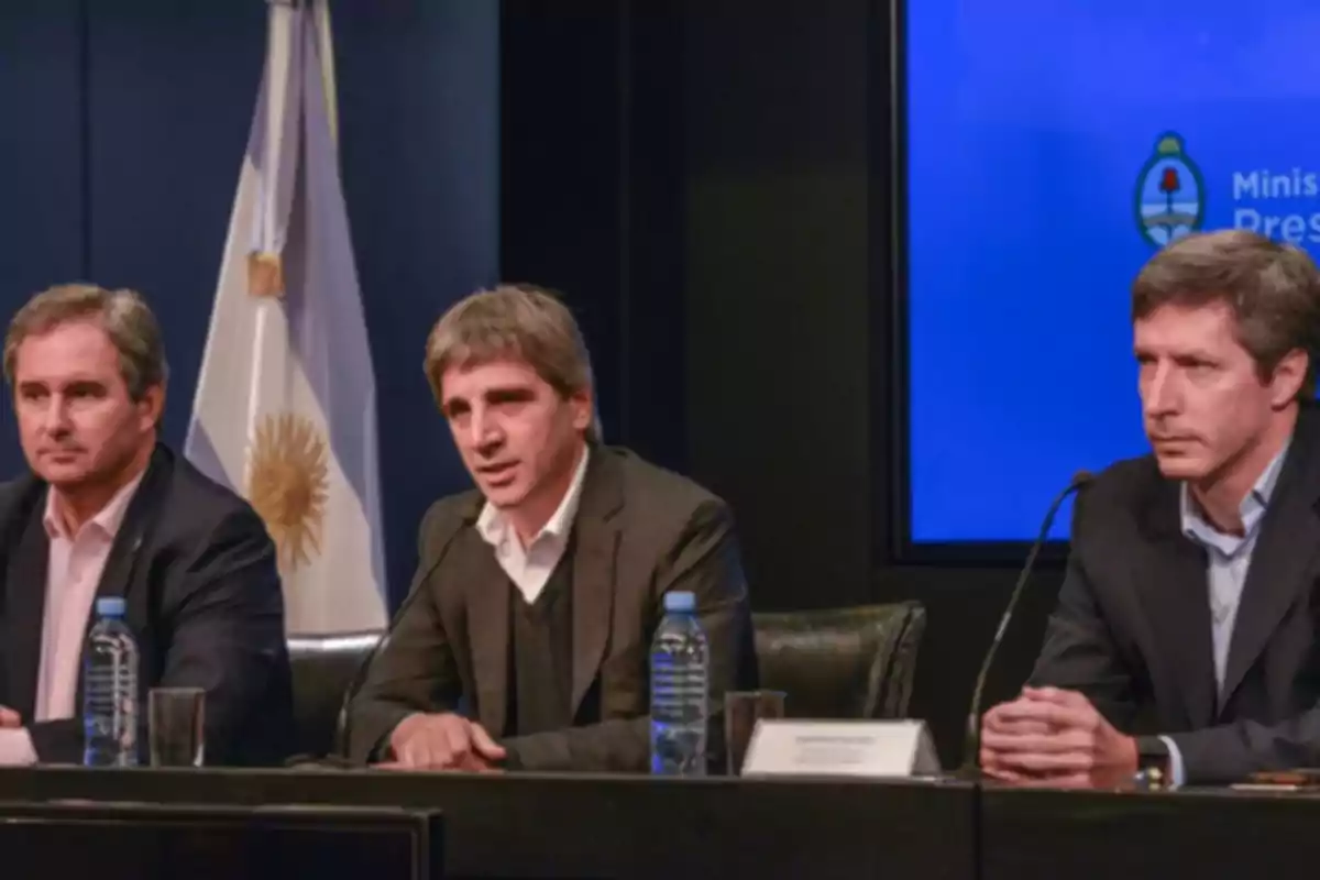 Tres hombres con traje sentados en una mesa durante una conferencia de prensa con una bandera de Argentina y una pantalla azul de fondo