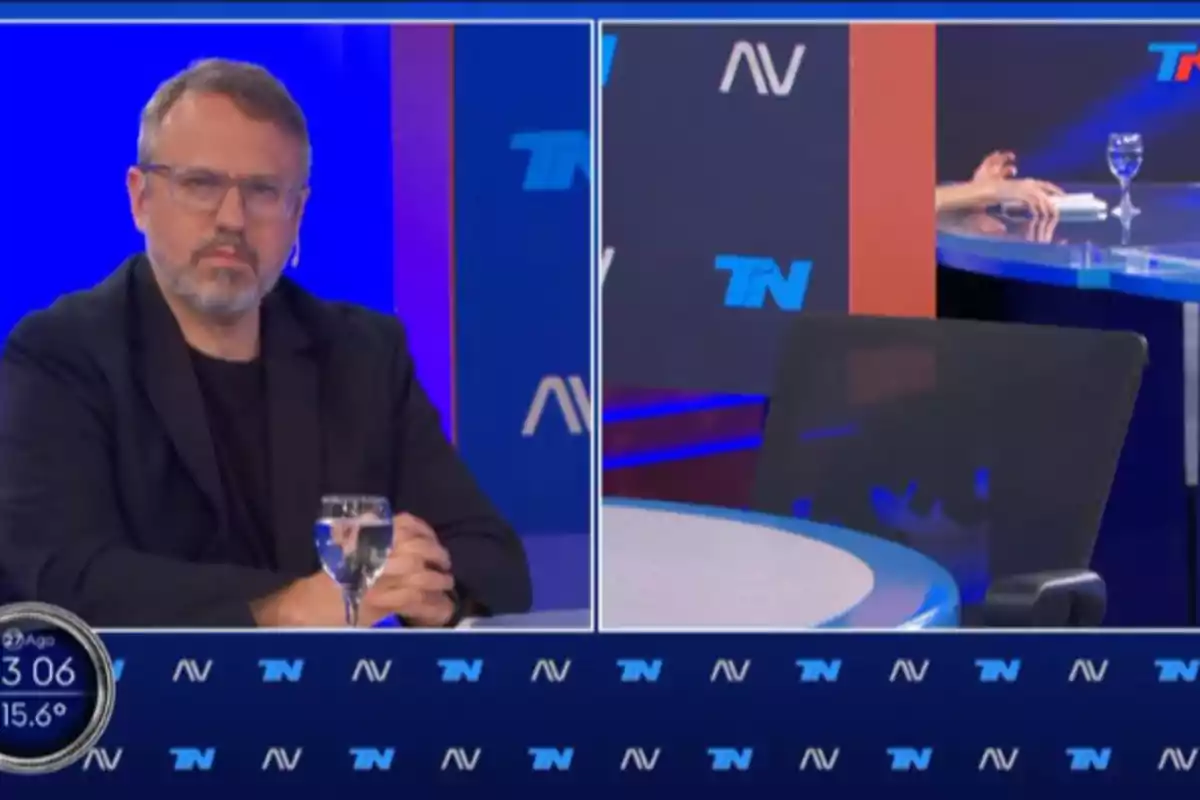 A man with glasses and a beard is sitting in a television studio with a blue background, while the chair next to him is empty and there are glasses of water on the table.